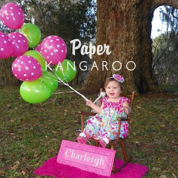 A young child sits in a rocking chair outdoors in front of a large tree, holding a bunch of pink and green balloons. A bright pink blanket on the ground holds a personalized sign reading 'Charleigh', and the words 'Paper Kangaroo' are overlaid across the center.