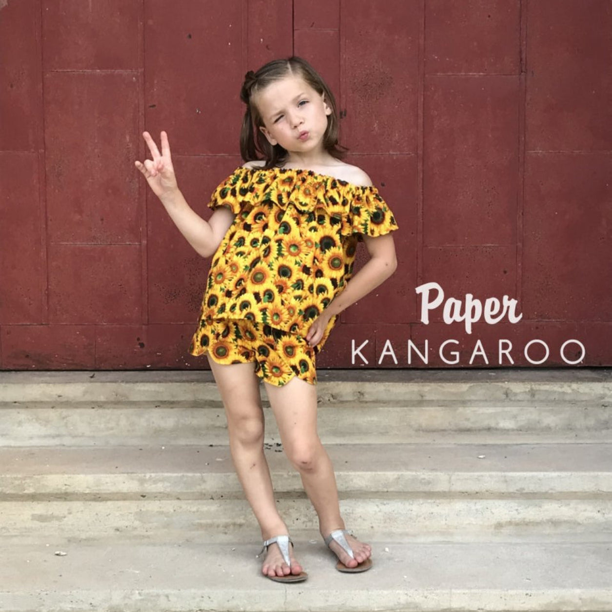 A young girl posing on concrete steps in front of a red wooden wall, wearing a yellow and black sunflower print off-the-shoulder romper. The text 'Paper KANGAROO' is visible on the right side of the image.
