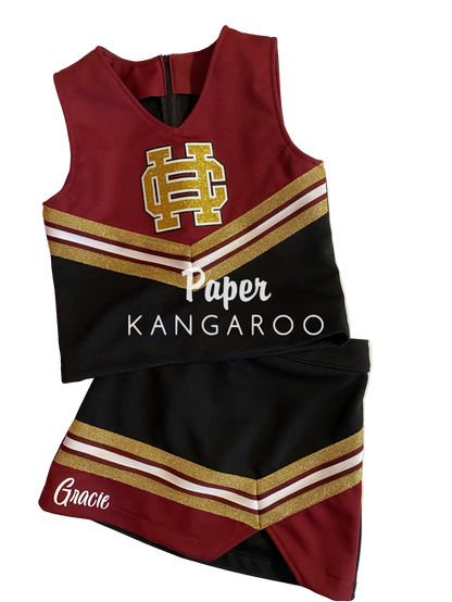 A cheerleader-style sleeveless top and matching skirt set laid flat on a white board. The outfit is primarily deep burgundy and black with gold glitter-style stripes and white trim.