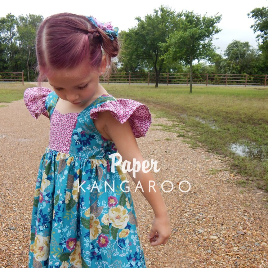 A young girl wearing a blue floral dress with pink patterned ruffle sleeves stands on a gravel path outdoors. The dress has a blue and green floral print with pink patterned shoulder ruffles and a pink patterned bodice.