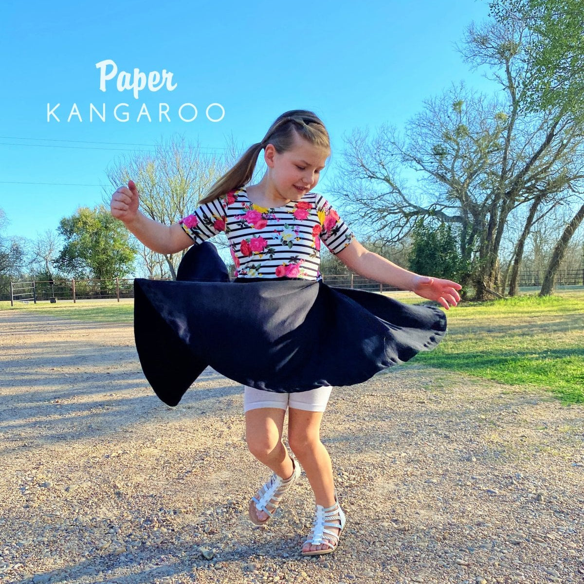 A young girl outdoors in a park wearing a black paper kangaroo skirt and a colorful striped shirt, spinning to show the black fabric flying out. The text 'Paper KANGAROO' is visible in the top left corner.