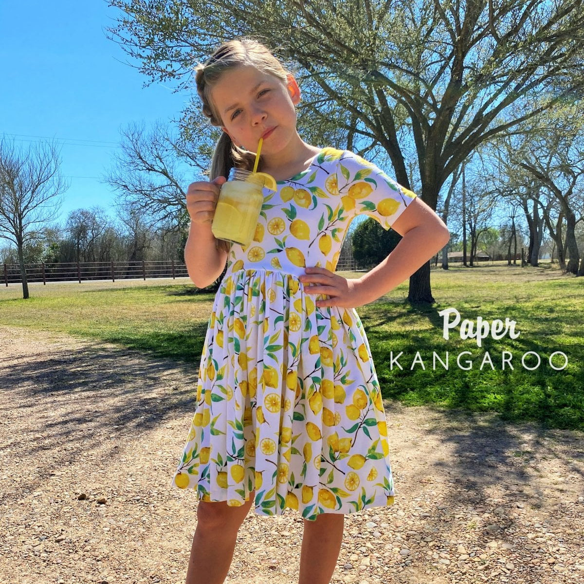 A young girl stands outdoors in a park-like setting wearing a white short-sleeve dress with a yellow lemon print. She is holding a yellow drink with a straw.