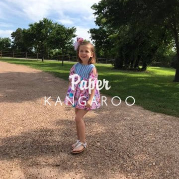 A young girl stands on a dirt path outdoors wearing a colorful patterned dress with a blue and white striped bodice and a pink floral skirt. Trees and grass are visible in the background, and a large text overlay reads 'Paper KANGAROO'.
