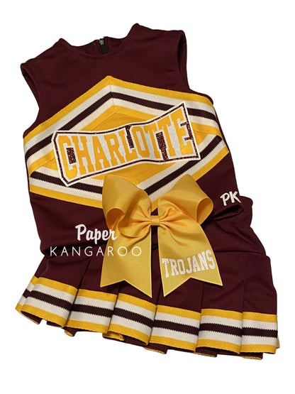 A toddler cheerleader dress in maroon, gold, and white, featuring a large gold bow at the waist, a pleated skirt, and 'CHARLOTTE TROJANS' printed on the front.