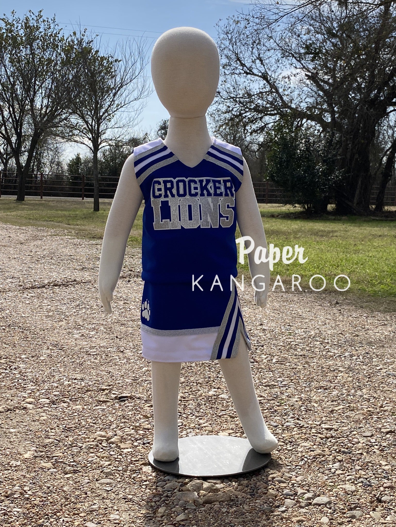 A toddler girl's cheer uniforms displayed on a white mannequin outdoors on a gravel path. The uniform is royal blue with white and silver stripes on the shoulders and sides, and 'CROCKER LIONS' printed in white across the chest.