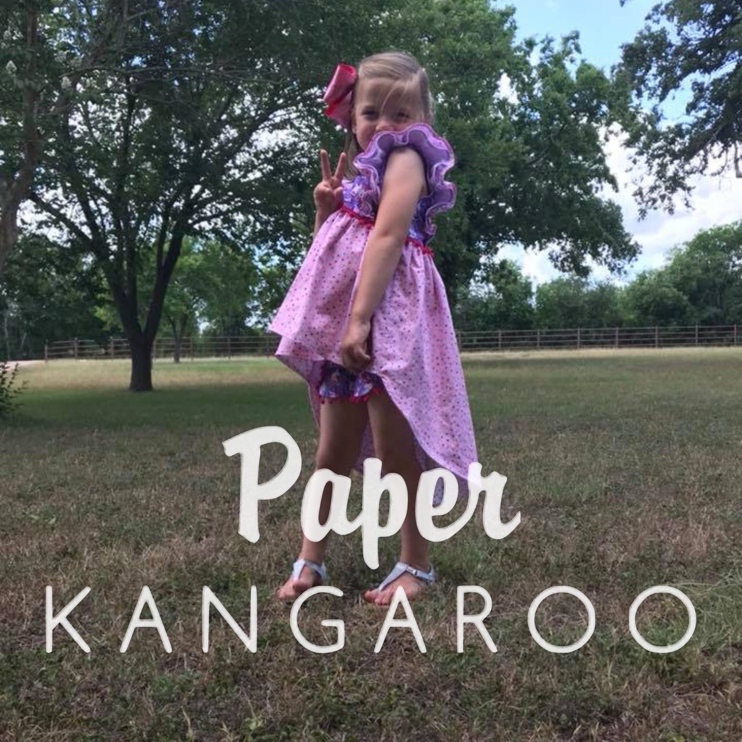 A young girl wearing a pink and purple patterned dress stands in a grassy outdoor field with trees in the background. Large white text overlay reads 'Paper KANGAROO'.