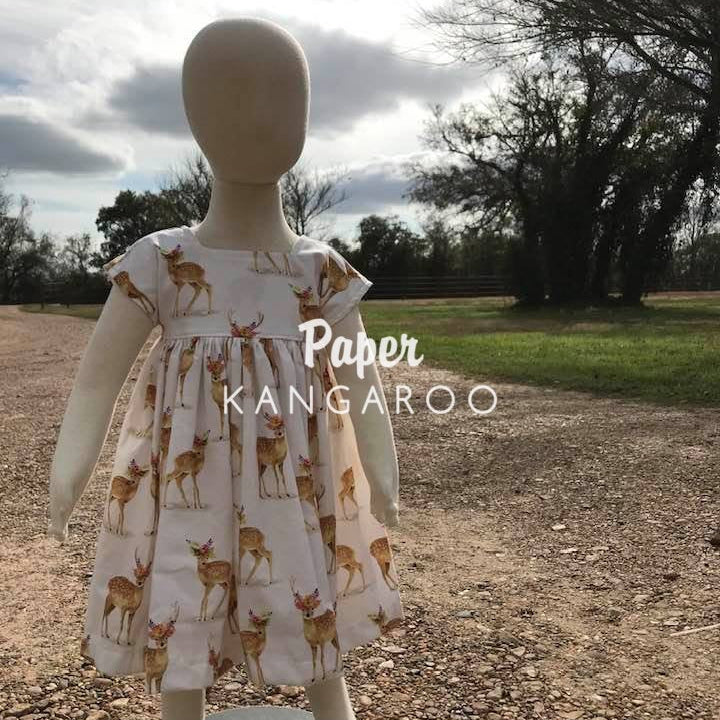 A child-sized white dress with a brown and tan deer print displayed on a mannequin outdoors on a gravel path with trees and grass in the background. A watermark reading 'Paper KANGAROO' is overlaid across the center.
