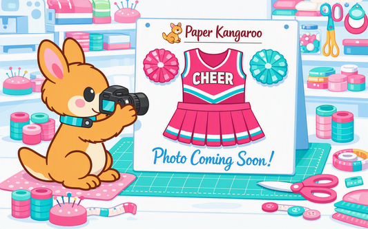 A stylized illustration of a cartoon kangaroo holding a camera beside a sign that says 'Paper Kangaroo' and 'Photo Coming Soon!' with a pink cheerleader dress design. The scene is a colorful craft room with spools of thread, scissors, and paper supplies.