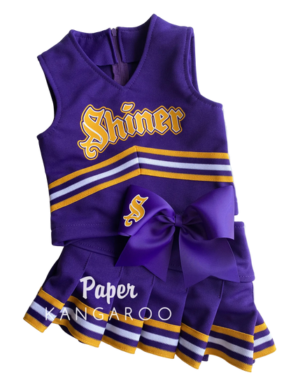 A purple girls' cheerleader costume set with a sleeveless top and matching pleated skirt, featuring gold and white stripes, a large purple bow, and 'Shiner' lettering on the front, photographed on a white background.