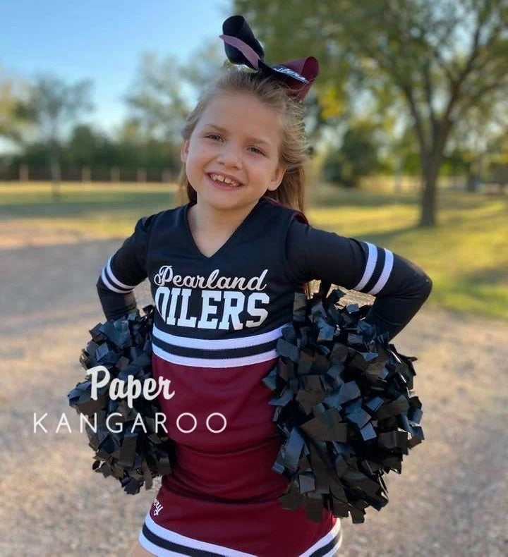 A young girl cheerleader standing outdoors in a park, wearing a black and maroon cheer dress with 'Pearland OILERS' text, a matching hair bow, and holding black pom-poms. A watermark reads 'Paper KANGAROO'.