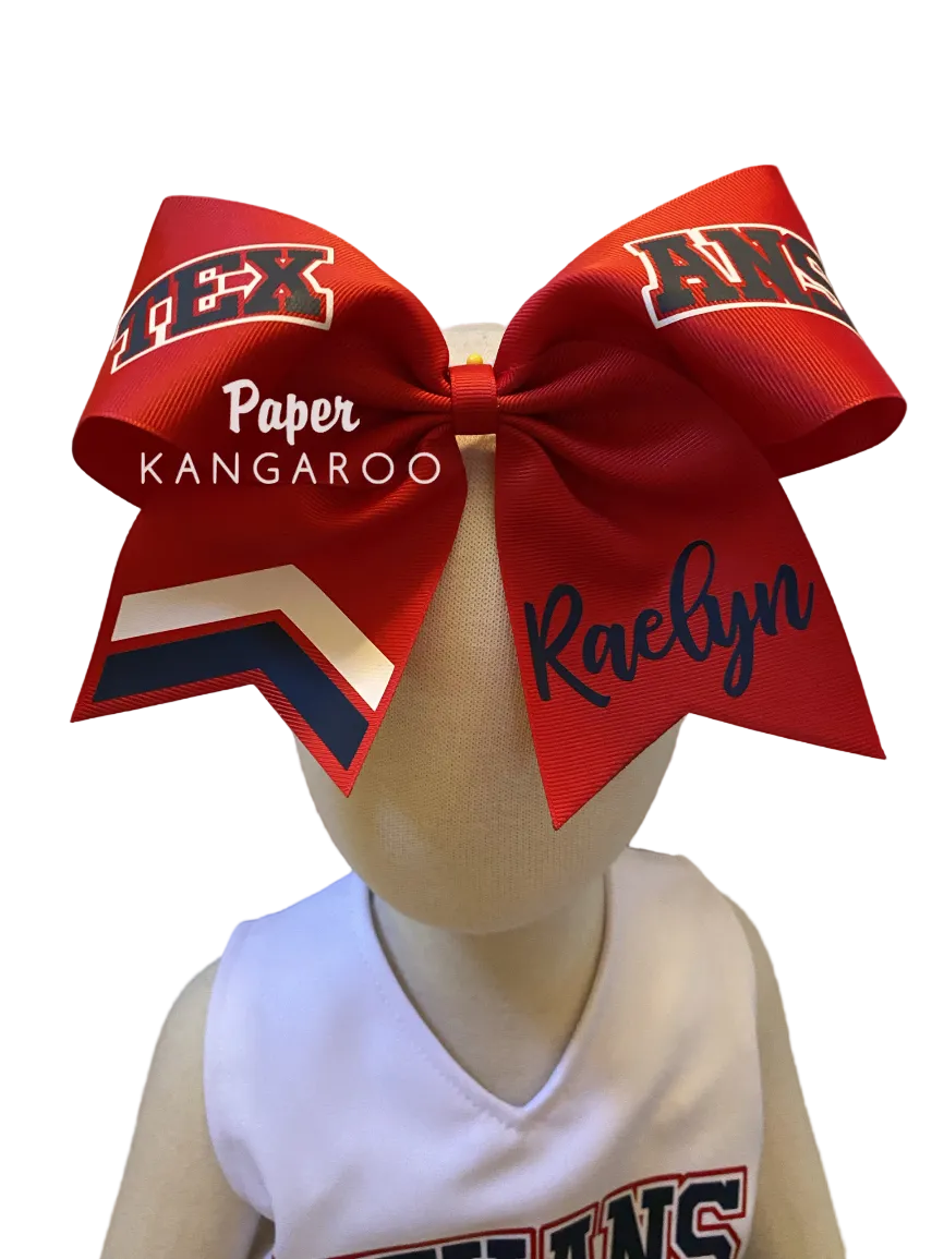 A large red cheerleader hair bow with navy and white lettering, displayed on a beige mannequin head wearing a white jersey. The bow features 'TEXANS' text and the name 'Raelynn' written in navy script.