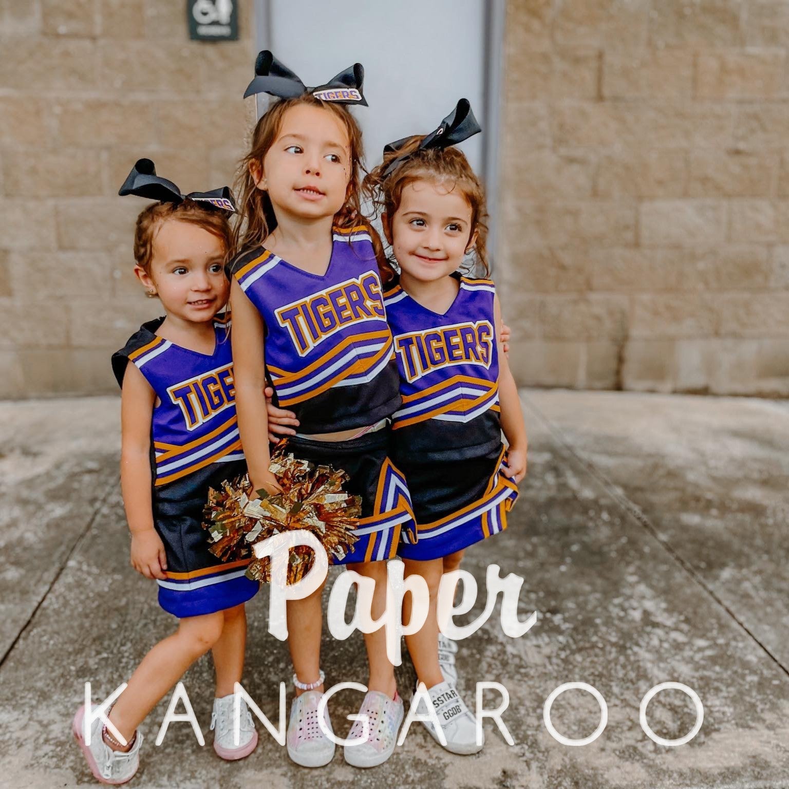 Three young girls wearing matching purple, black, and gold cheerleader outfits with 'TIGERS' lettering, standing outdoors in front of a tan brick wall. Large white text overlay reads 'Paper KANGAROO'.