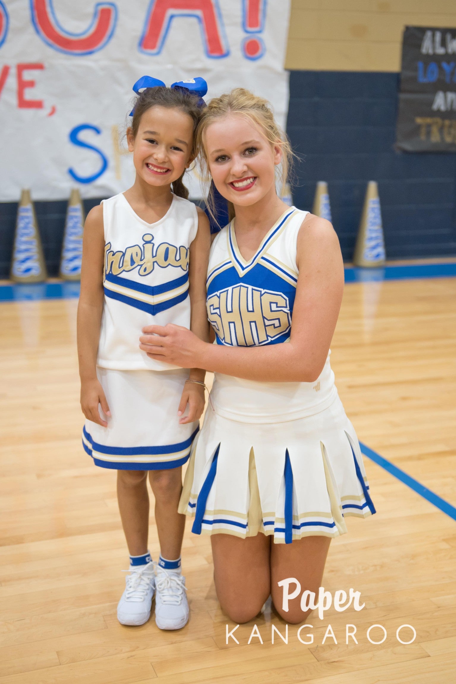 A woman and a young girl wearing matching white and blue cheerleader uniforms pose together on a school gymnasium basketball court. The woman is kneeling beside the girl, and both have 'SHS' lettering on their outfits.