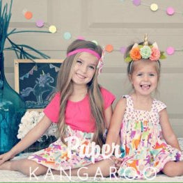 Two young girls sitting side-by-side wearing colorful floral headbands and matching floral dresses, with a 'Paper Kangaroo' text overlay at the bottom.