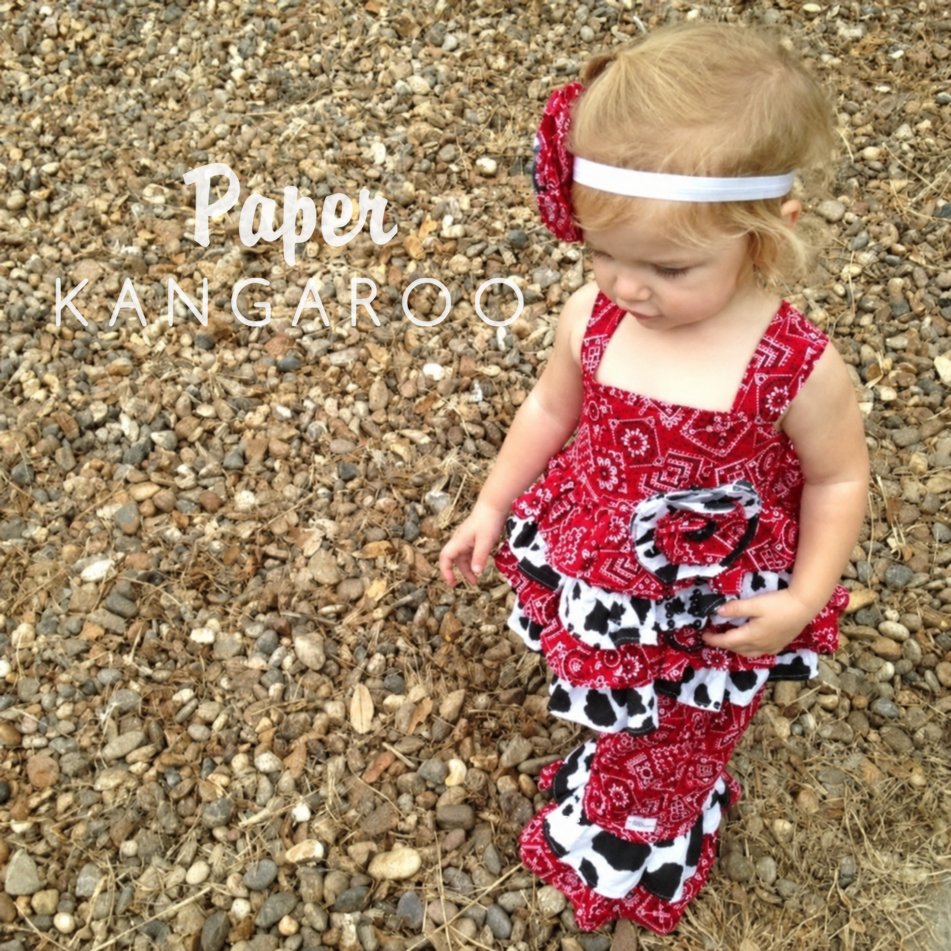 A toddler stands outdoors on a bed of small brown and gray pebbles wearing a red, black, and white cow print dress with a matching headband. White text overlay on the left reads 'Paper KANGAROO'.