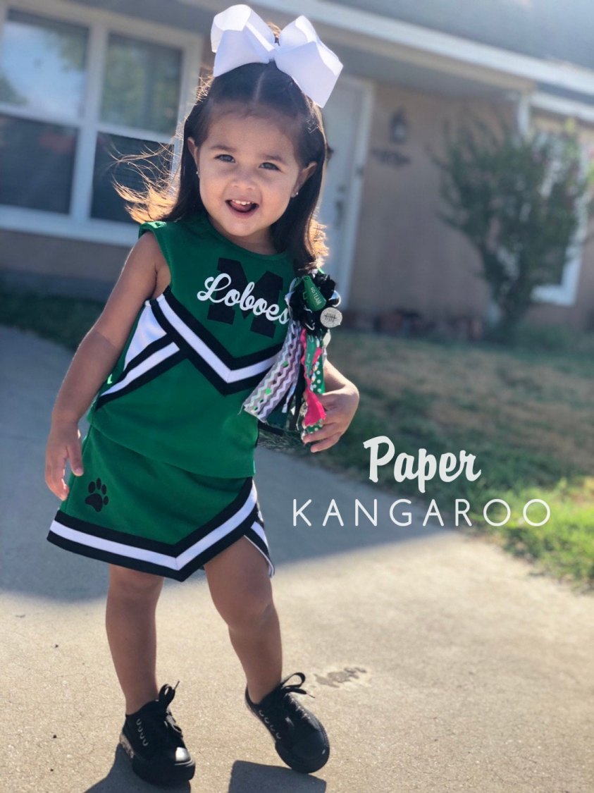 Toddler girl wearing custom green and black Loboes cheer uniform with paw print skirt detail, white hair bow, and spirit mum — handmade by Paper Kangaroo