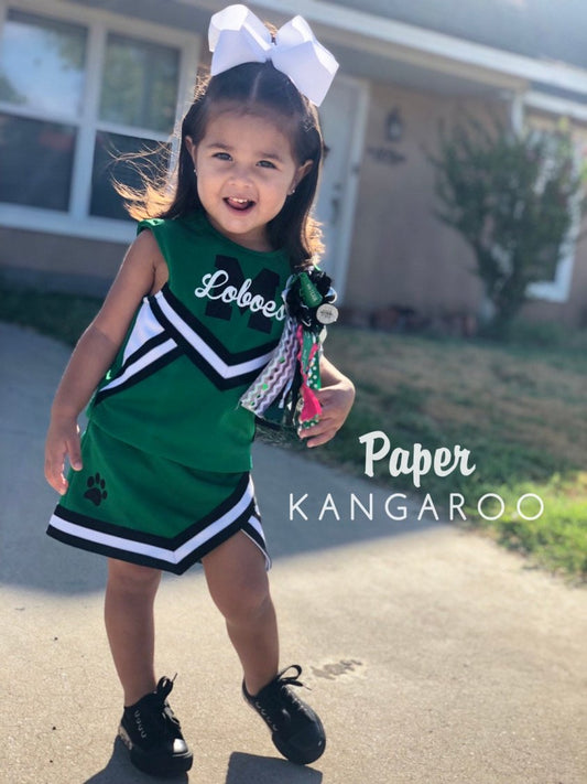 Toddler girl wearing custom green and black Loboes cheer uniform with paw print skirt detail, white hair bow, and spirit mum — handmade by Paper Kangaroo